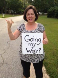 Audrey on the side of the road with her thumb out and a sign that reads "Going My Way?"